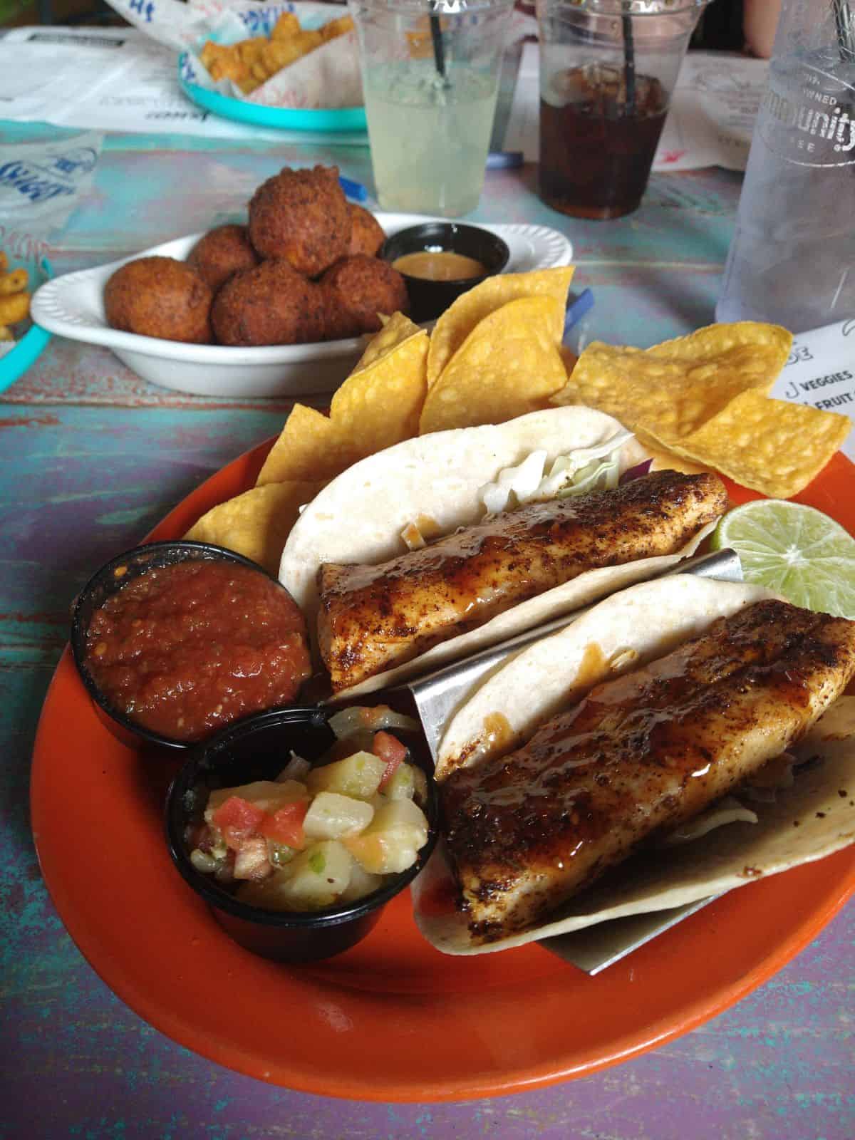 Mahi Mahi fish tacos on a red plate at a restaurant serving with chips, salsa, cocktail sauce, and hushpuppies. 