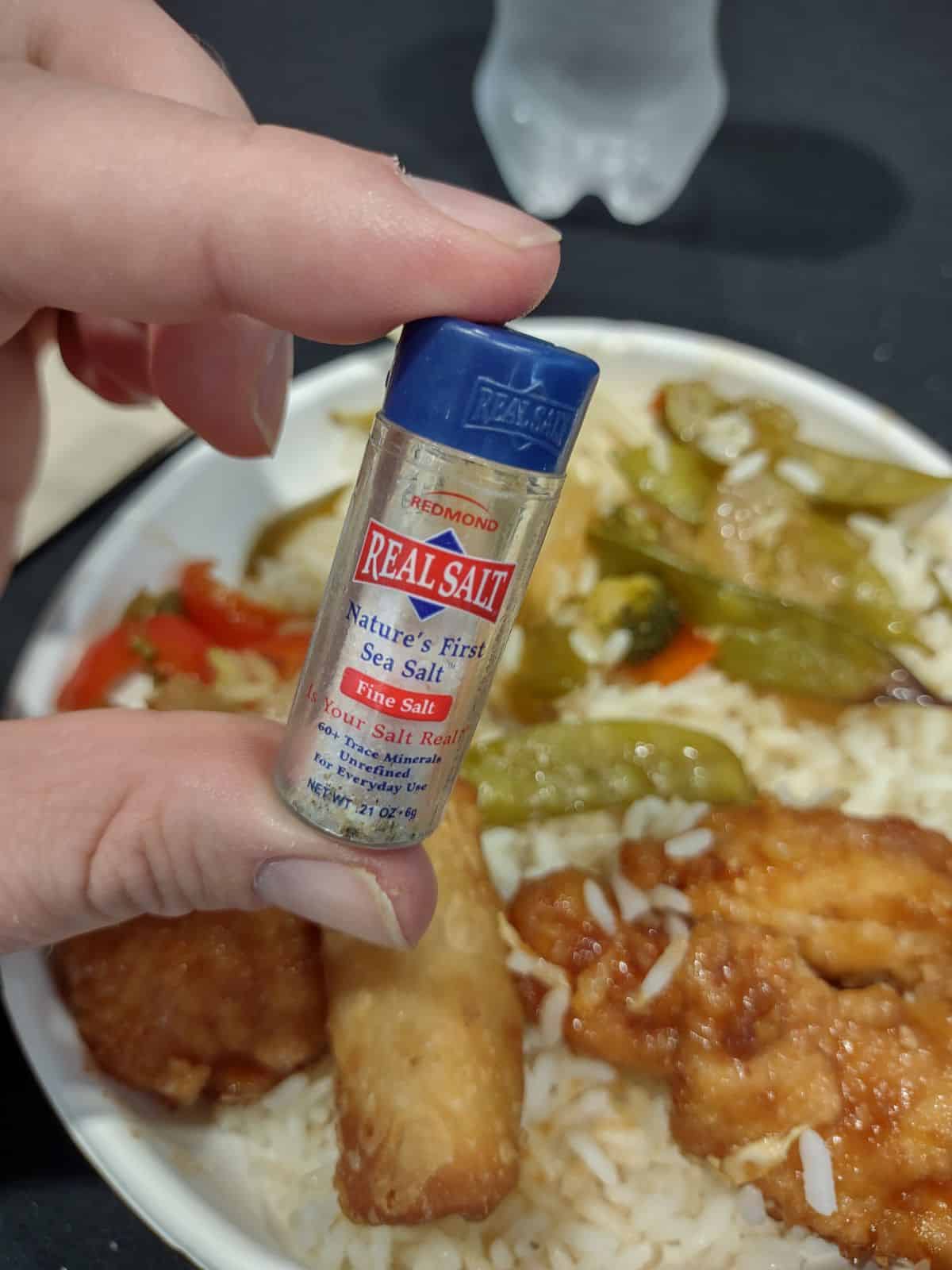 Holding a mini Redmond Real Salt shaker over top a plate of Chinese food with chicken, rice, and veggies.