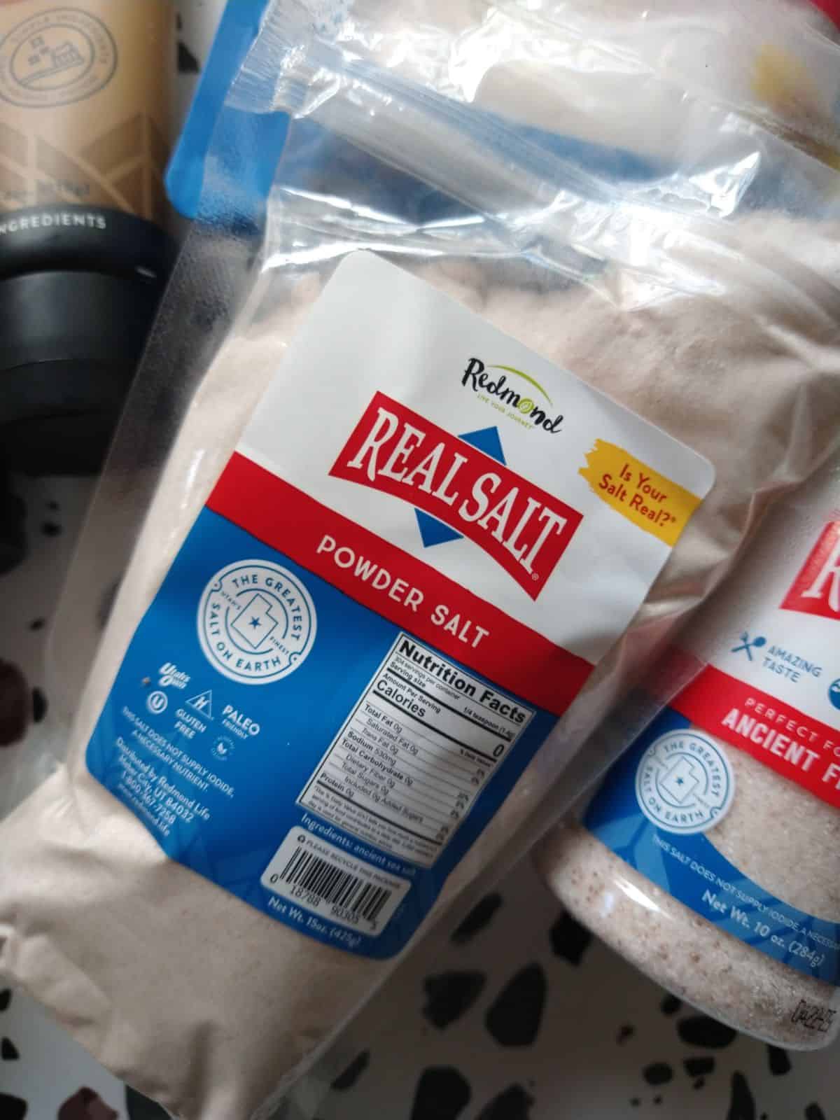 A bag of Redmond Real Salt Powder Salt sitting next to a Redmond Real Salt shaker.