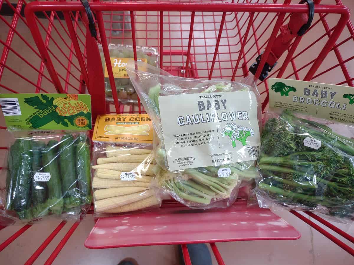 Bags of baby zucchini, baby corn, baby cauliflower, and baby broccoli in a red Trader Joe's shopping cart.