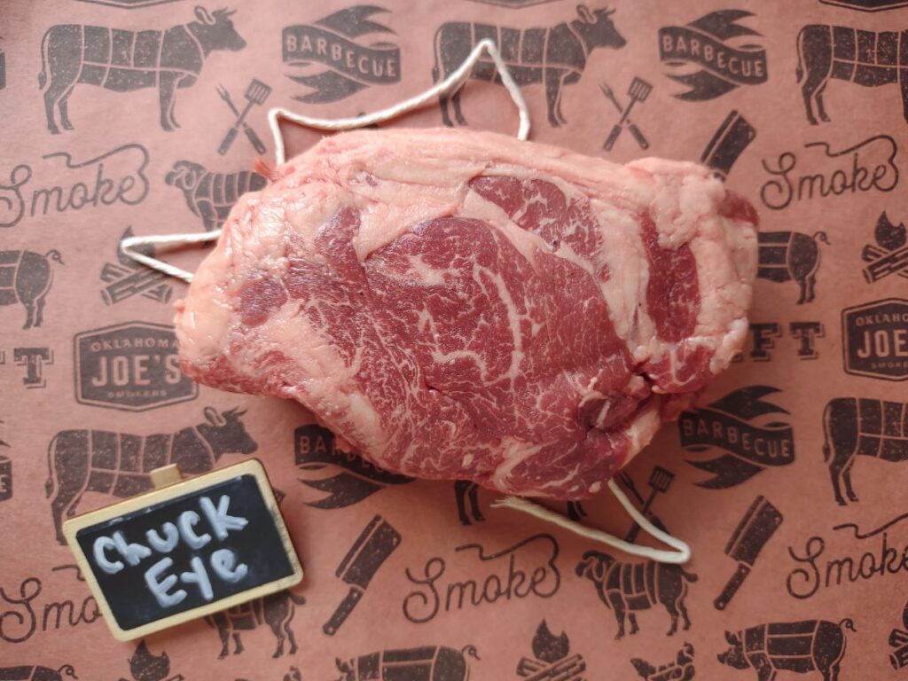 A raw chuck eye steak sitting on a piece of butcher paper with a string underneath it and a small chalkboard sign that says "chuck eye".