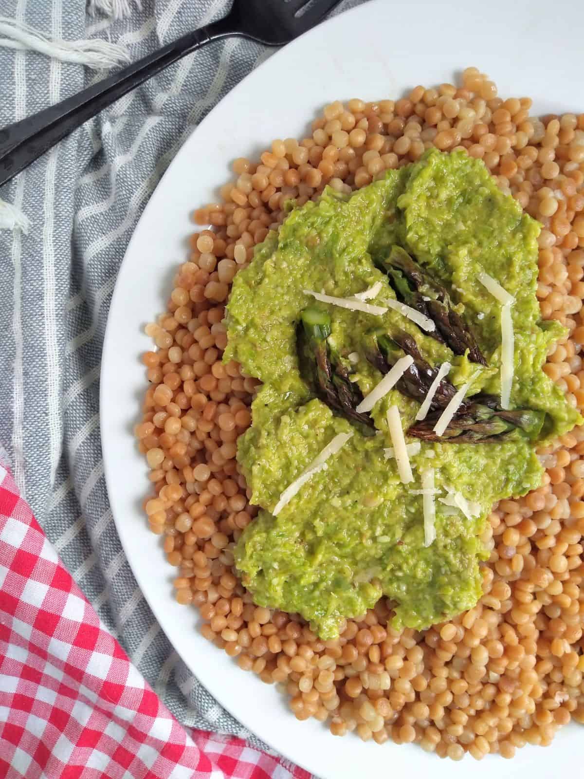 Toasted couscous topped with asparagus pesto, asparagus tips, and parmesan cheese on a white plate. 