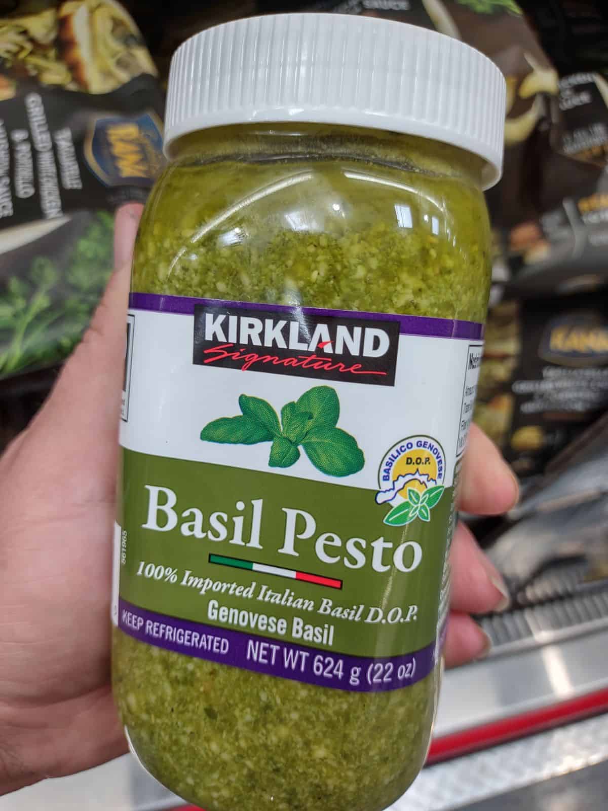 Holding up a jar of Kirkland Brand Basil Pesto with 100% Imported Italian Basil DOP.
