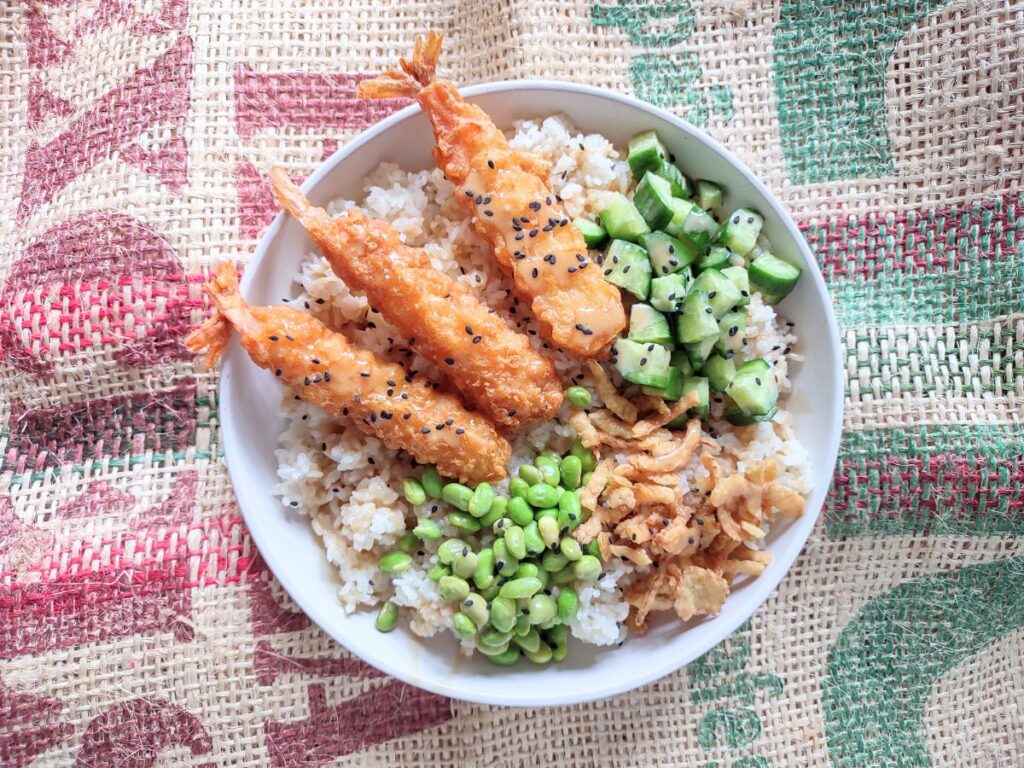A rice bowl that has been toppped with tempura shrimp, edamame, crispy onions, and cucumber.