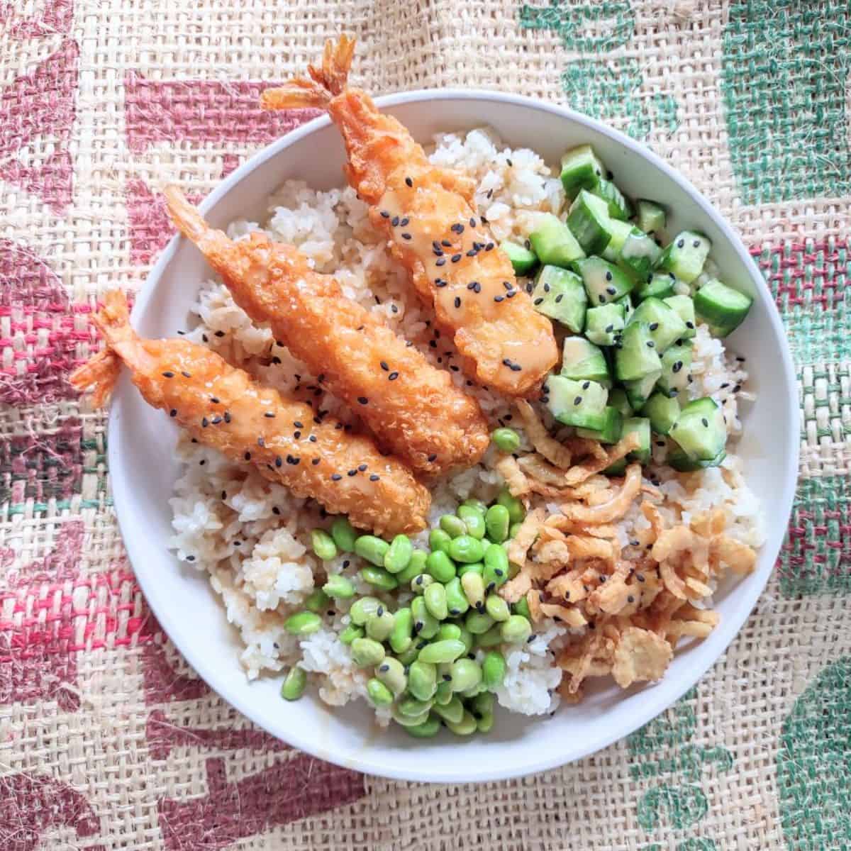 A bowl of tempera shrimp with cucumbers, edamame, crispy onins, over white rice.