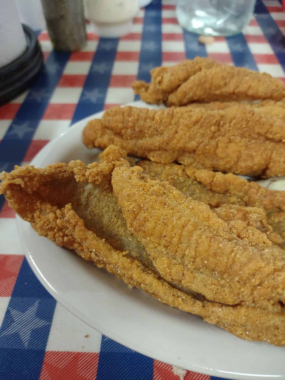 Breaded fried catfish on a white plate with an Americana table cloth.