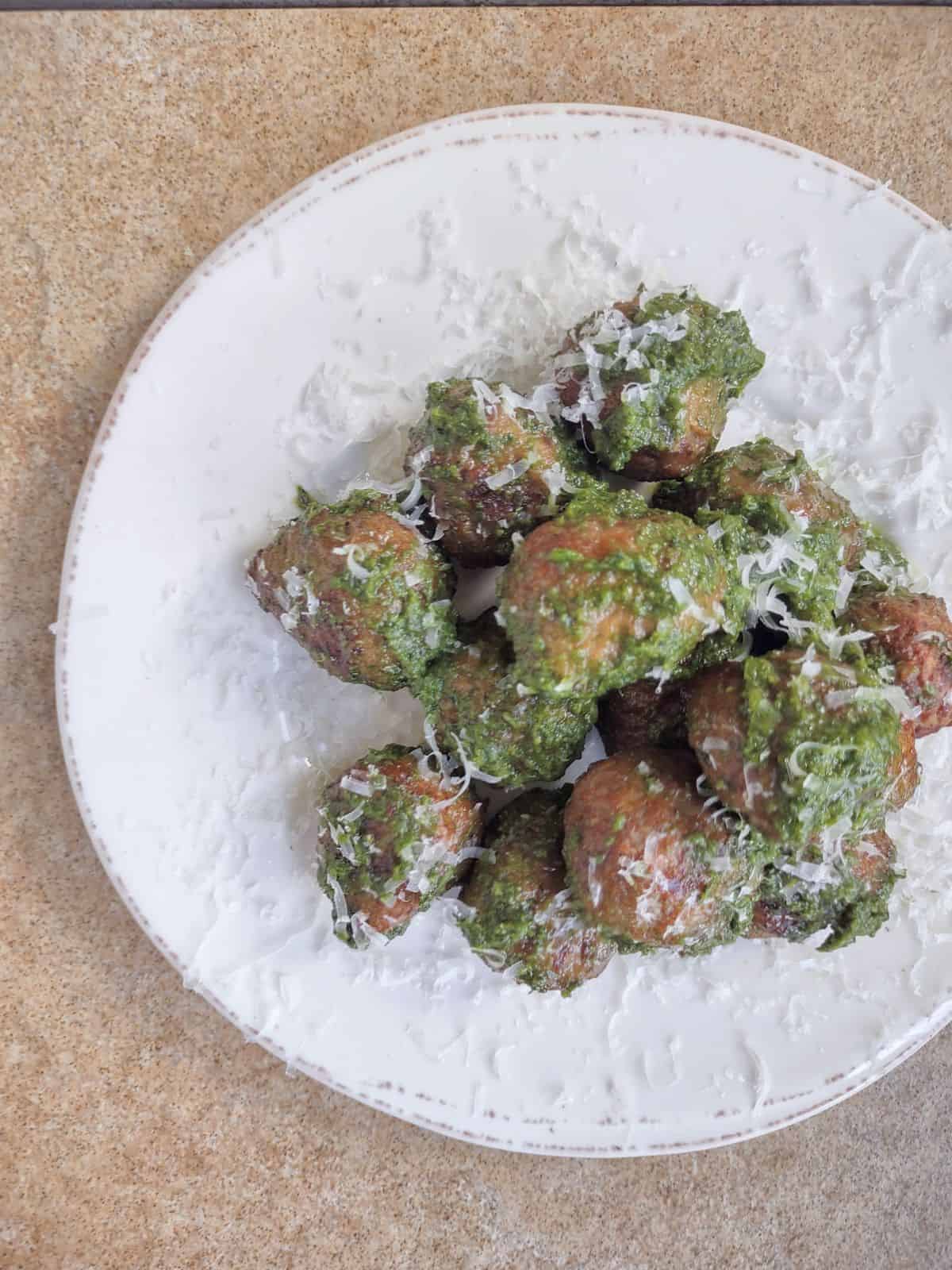 Cooked meatballs on a plate bowl covered in pesto, and grated parmesan cheese.