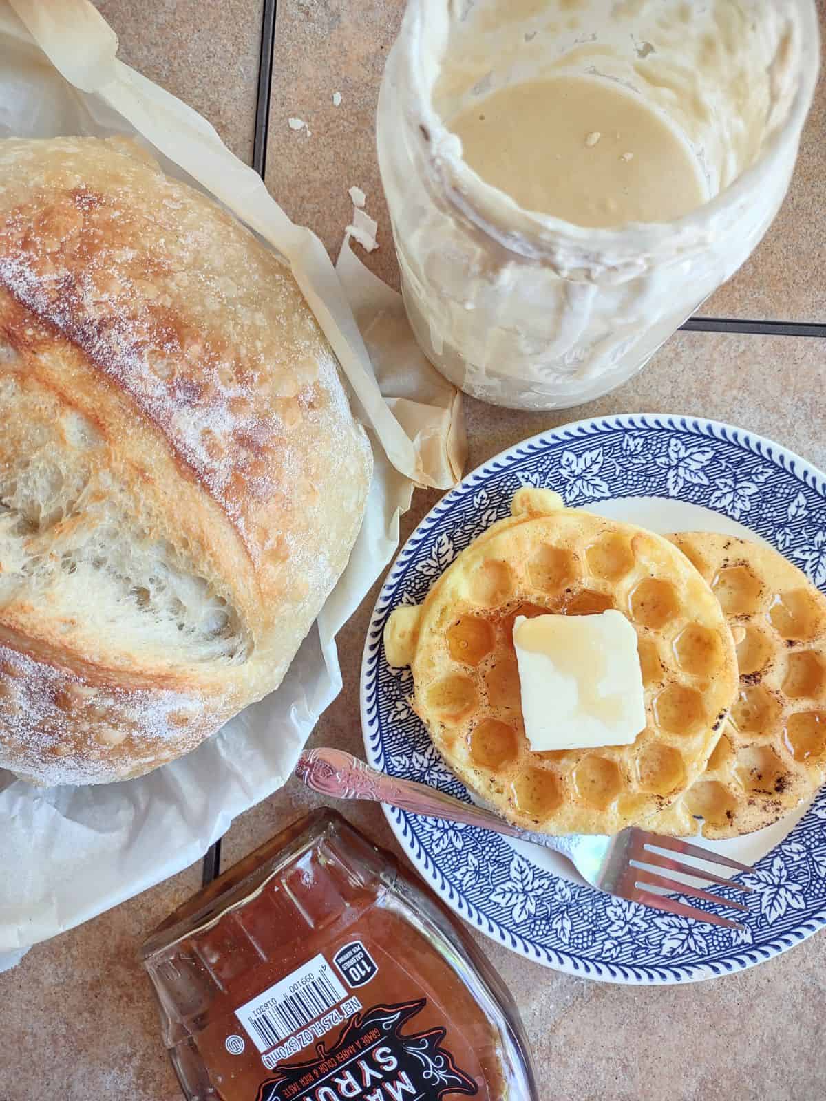 Sourdough discard waffles on a plate next to a loaf of sour dough with a jar of sourdough starter.