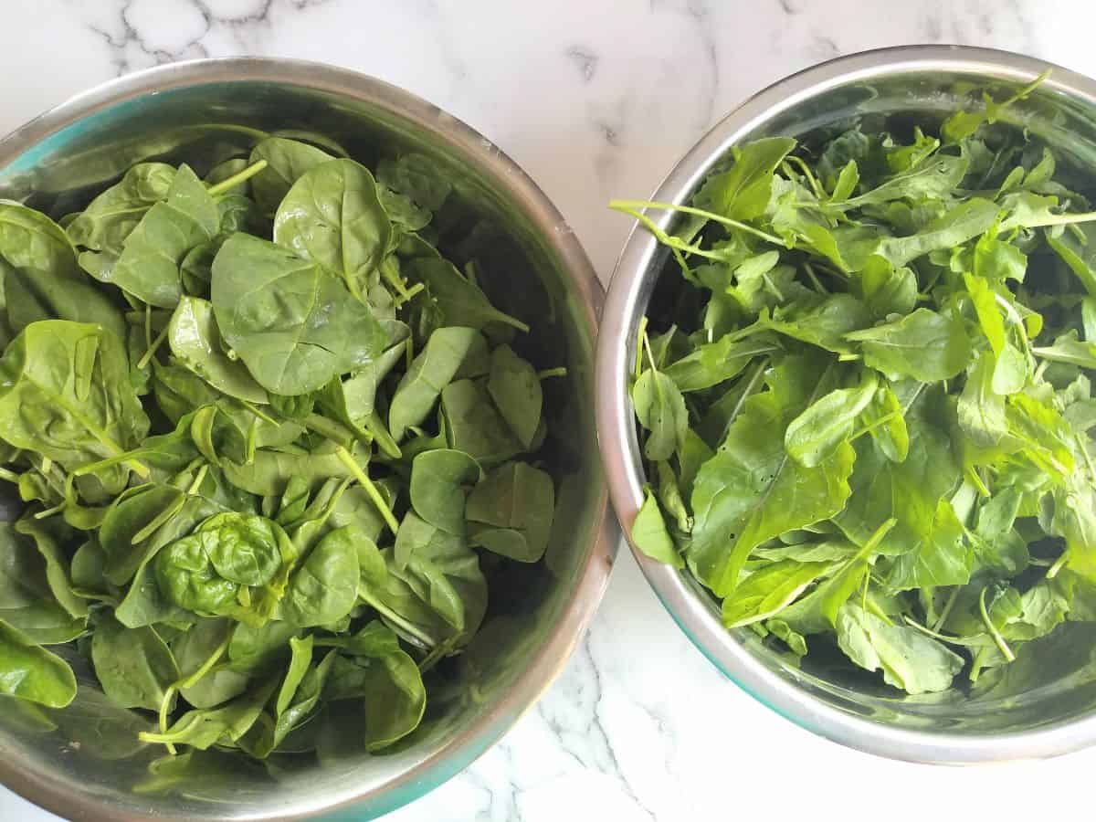 A metal bowl of baby spinach and a metal bowl of arugula.