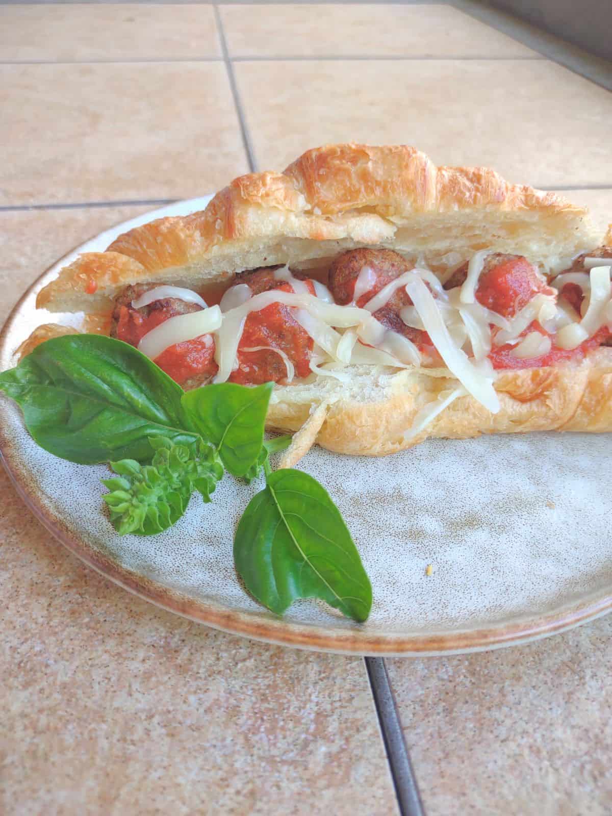 A plate with a croissant filled with cooked meatballs, mozzarella cheese, and tomato sauce.