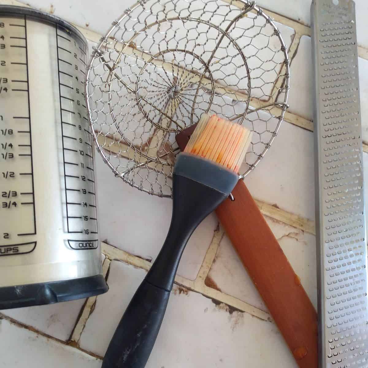 A plunger cup, Microplane zester, silicone brush and spider sit on a white tile counterop.