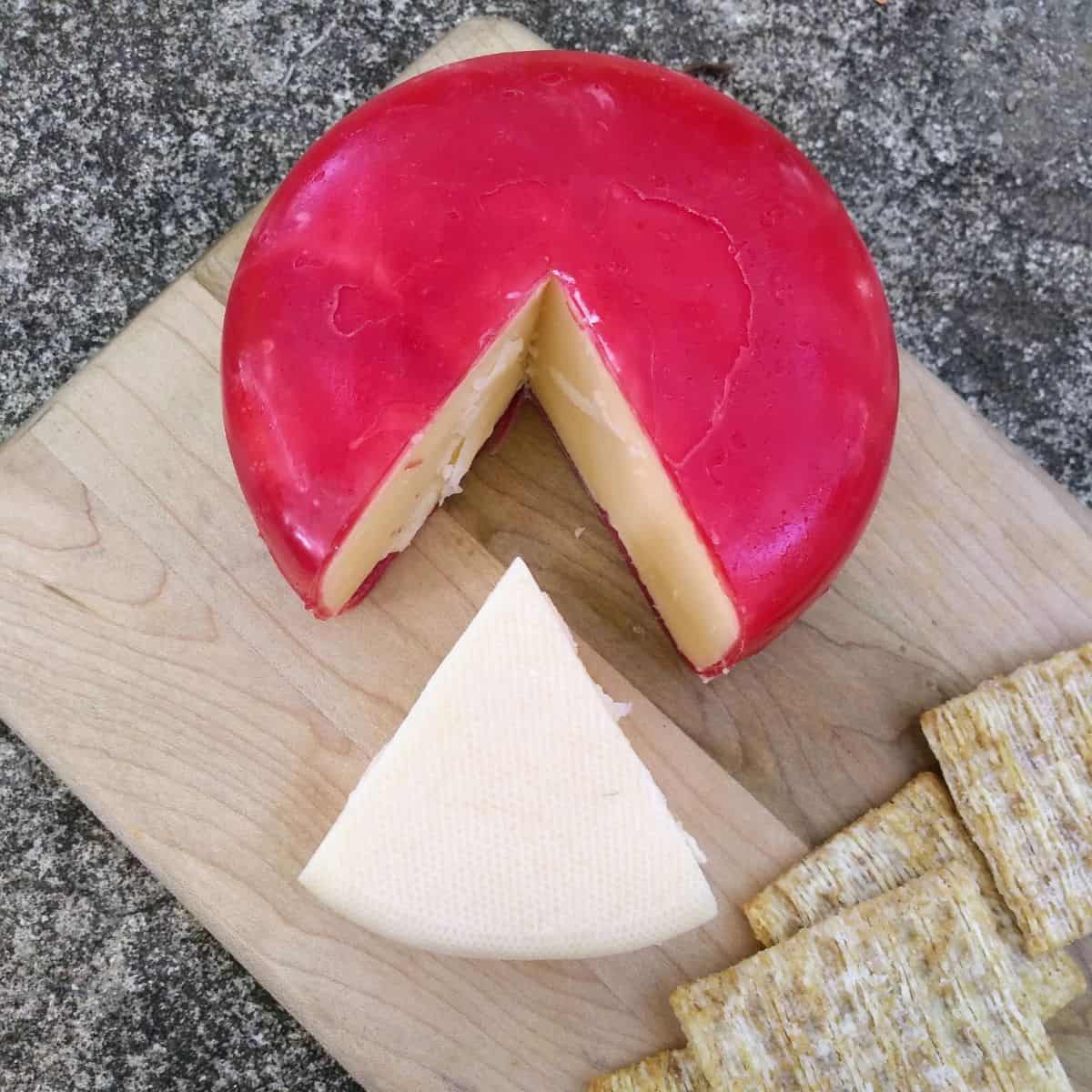 A piece of gouda cheese cut from a wheel of red waxes gouda, sitting on a cutting board with crackers.