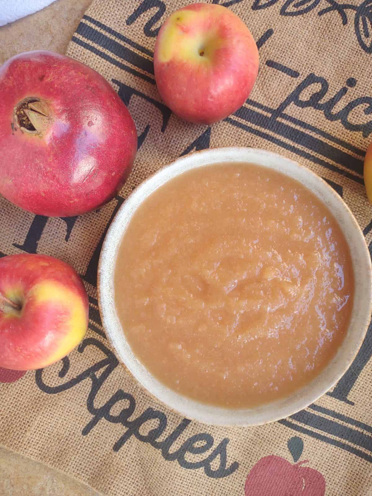 A bowl of homemade pomegranate applesauce with apples and pomegranates around the bowl.