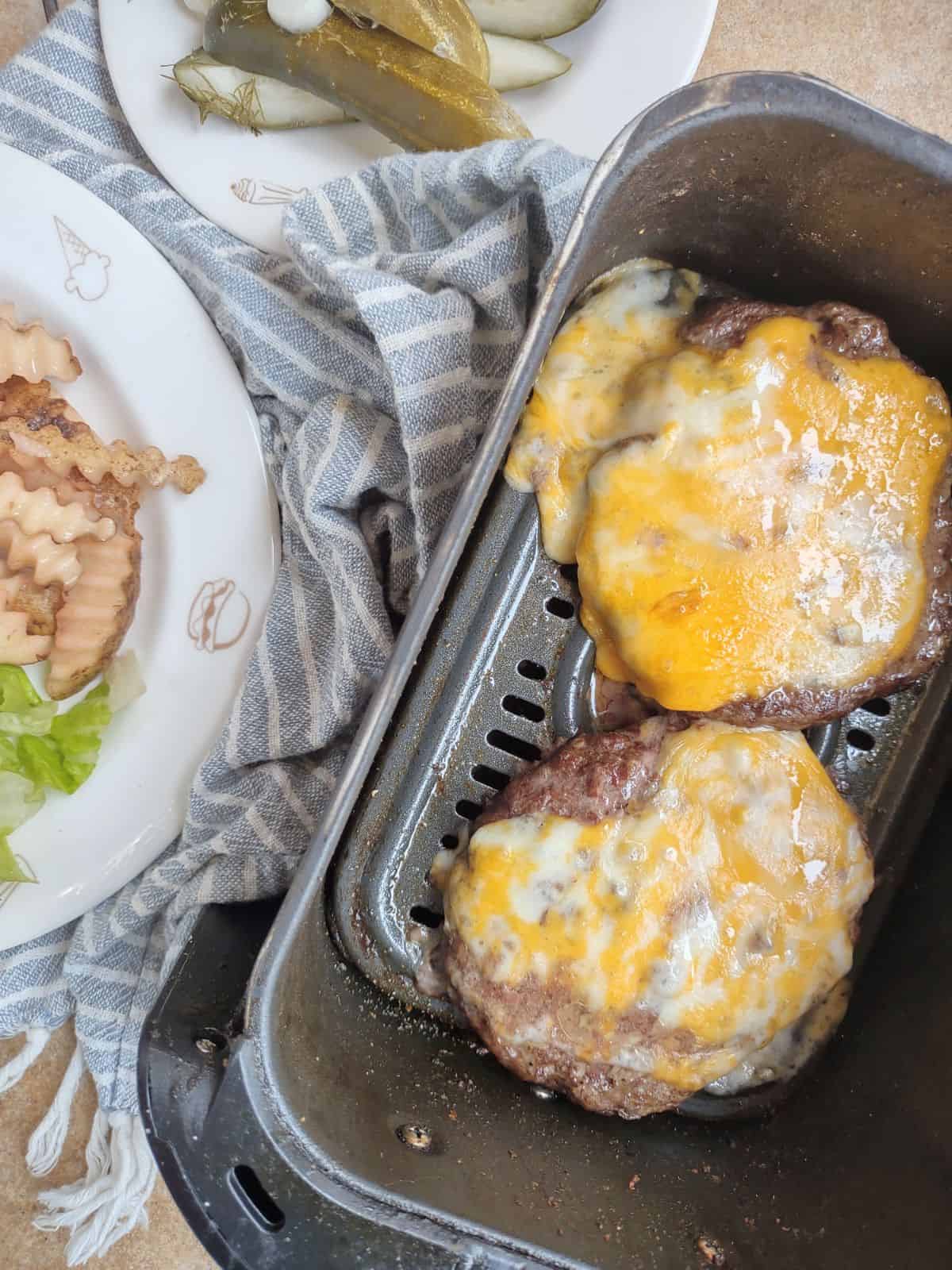 Cheese burgers in an air fryer basket with a plate of fries and lettuce next to it and a plate of pickles.