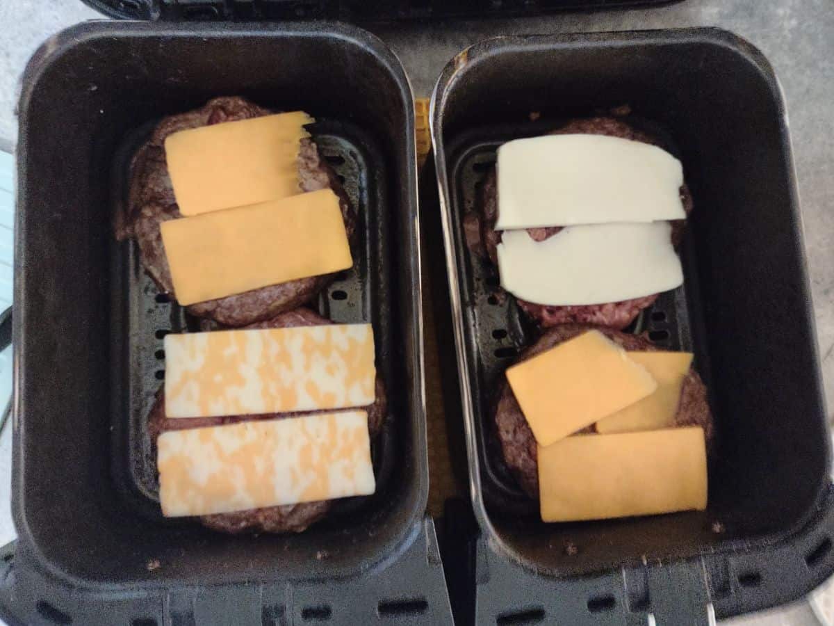 Cheese placed over top of partly cooked burger patties in two air fryer baskets.