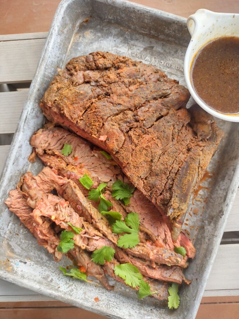 A smoked flank steak on a sheet pan, with pieces sliced off, along with au jus and some chopped cilantro.