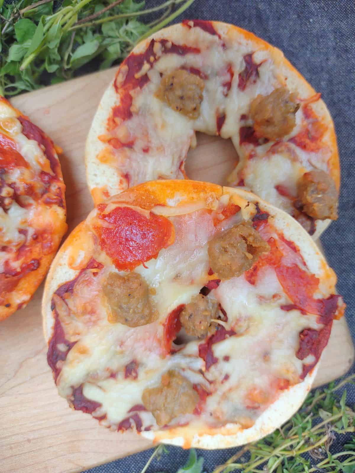 3 Sausage and pepperoni pizza bagels on a wood cutting board surrounded by herbs.