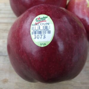 Up close view of a red Macoun apple on a wood board. The apple has a Fowler Farms stick on it.