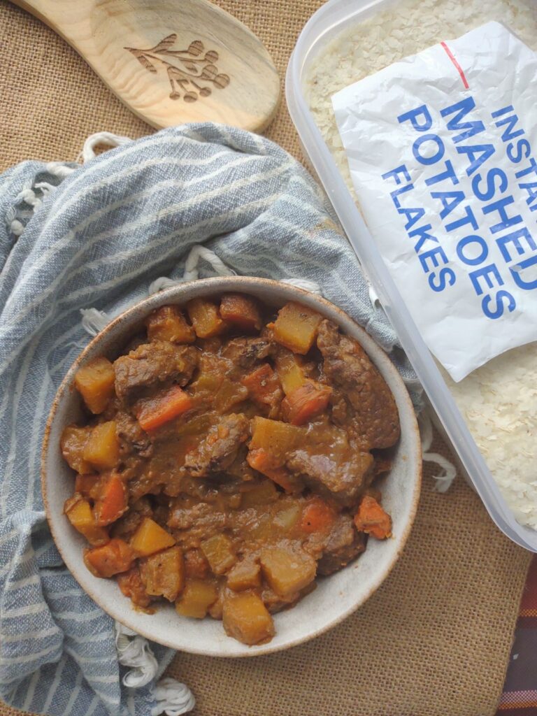 A bowl of beef stew with a container of mashed potato flakes next to it.