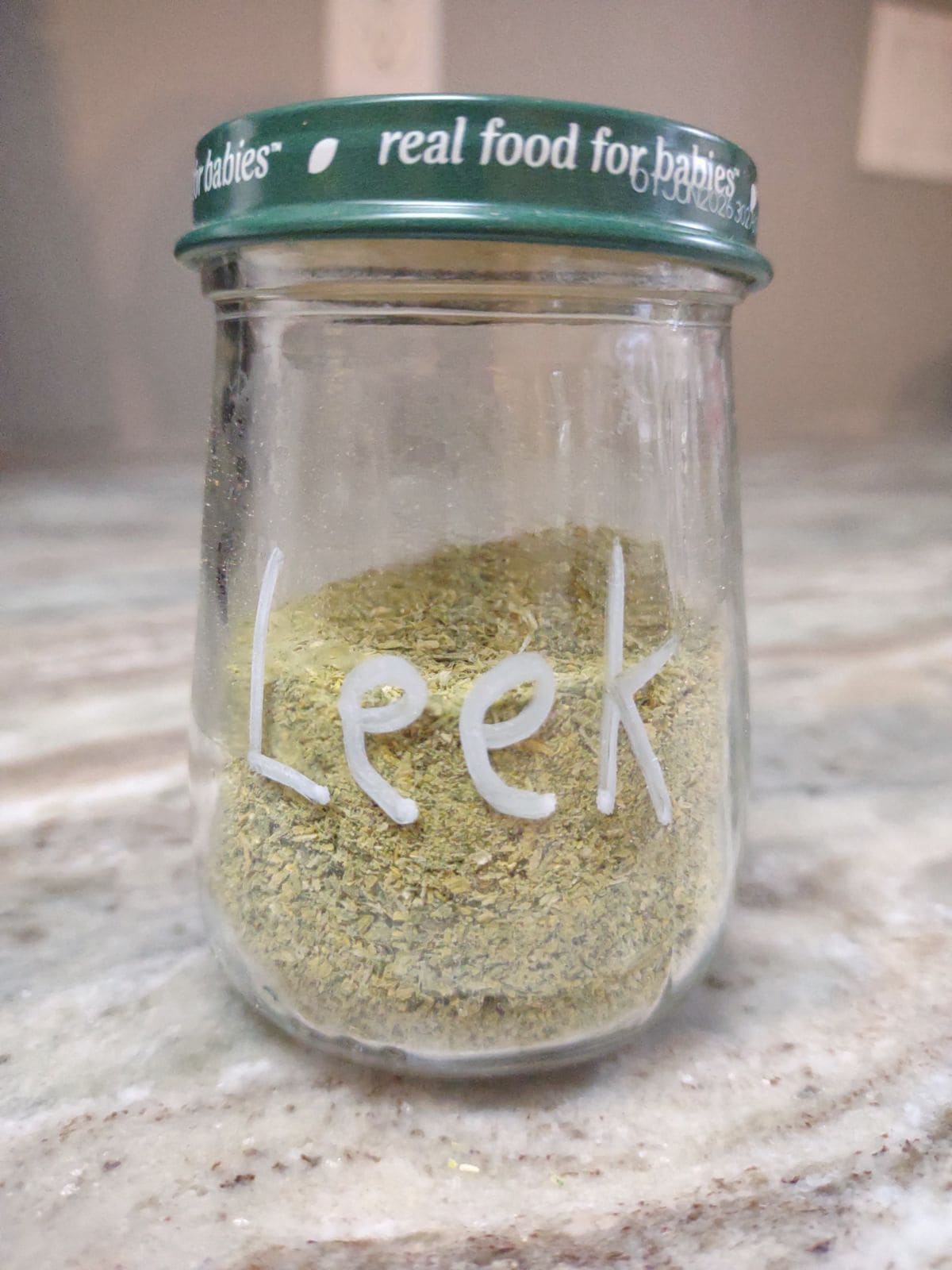 A small, clear glass jar with a lid that contains homemade green leek powder.