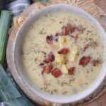 A bowl of Potato Leek soup that is topped with bacon and dried thyme on top of a plate with leeks next to the plate.