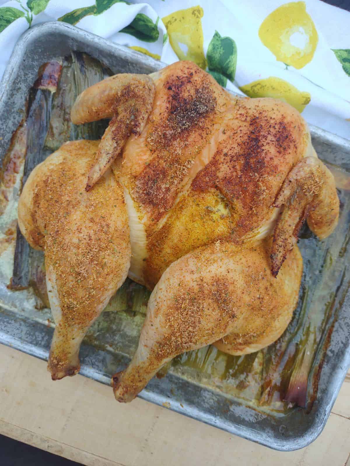 A whole butterflied roast chicken on top of the green parts of leeks on a sheet pan.