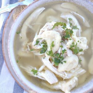 Chicken and dropped dumplings soup in a bowl on top of a plate on top of a blue and white stripe towel.