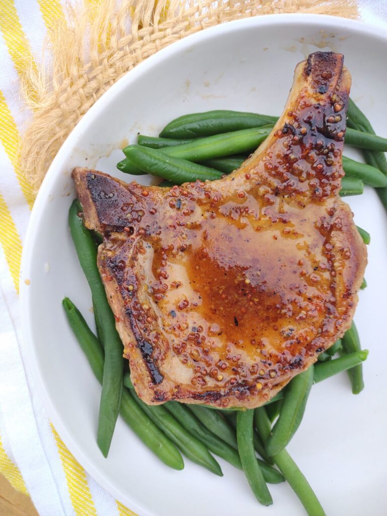 A pork chop that is glazed with a honey mustard sauce on top of some green beans on a white plate.