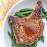 A pork chop that is glazed with a honey mustard sauce on top of some green beans on a white plate.