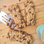 3 Tahini Chocolate Chip Granola bars on a wood cutting board with a metal spatula and a bottle of Tahini.