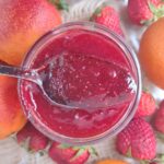 A spoon above a jar of Blood Orange Strawberry jam above a jar surrounded by strawberries and blood oranges.