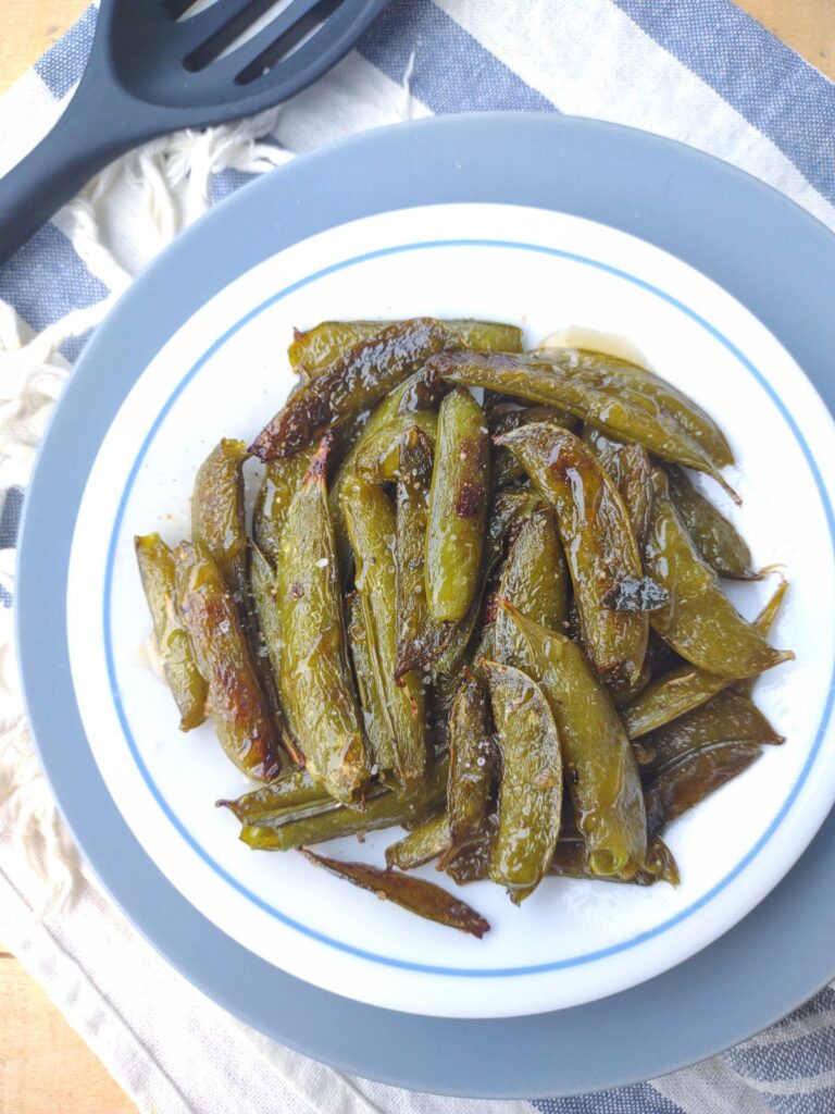 Roasted snap peas with honey on a white plate on top of a blue plate and white and blue stripe towel.