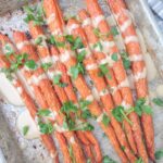 A sheet pan filled with roasted whole carrots that are topped with Tahini sauce and fresh parsley.