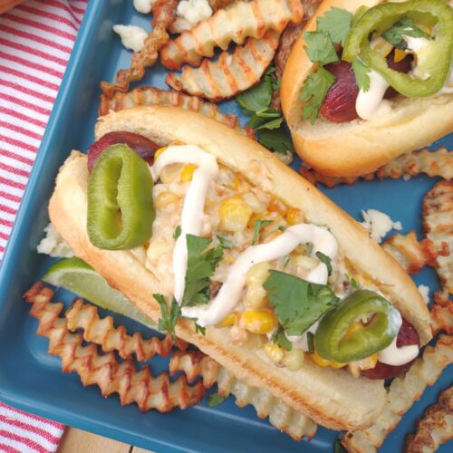 Hot dogs topped with Elote on a blue sheet pan with crinkle cut fries, lime wedges, cilantro, and cheese.