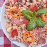 A pizza pasta salad with pasta, pepperoni, peppers, olives, and a sauce in a white bowl.