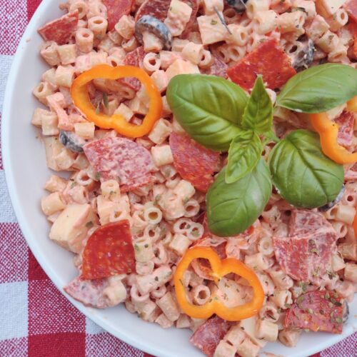 A pizza pasta salad with pasta, pepperoni, peppers, olives, and a sauce in a white bowl.