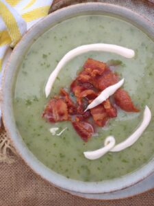 Soup made with asparagus ends, potatoes, and watercress in a bowl topped with crema and bacon.