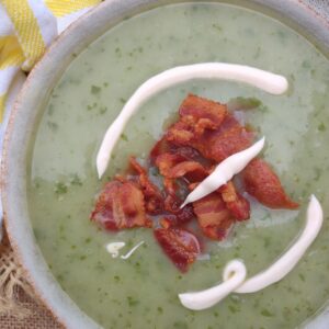 Soup made with asparagus ends, potatoes, and watercress in a bowl topped with crema and bacon.