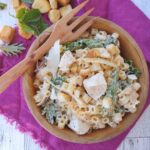 A wood bowl of pasta salad with Caesar dressing, baby kale, chicken, and parmesan.