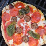An up close picture of a grilled pizza with pepperoni, sausage, and basil.