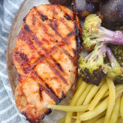 A grilled pork chop that has been coated in BBQ sauce with purple broccoli, purple potatoes, and wax beans on a plate.