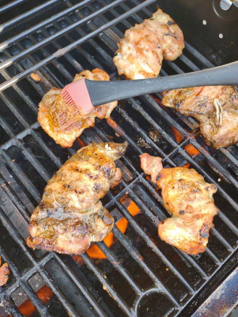 Chicken thighs on the grill with a brush that is brushing on a honey garlic and ginger sauce.
