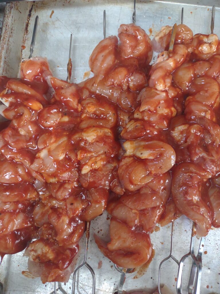 Raw chicken thigh meat on metal skewers with BBQ sauce brushed on.