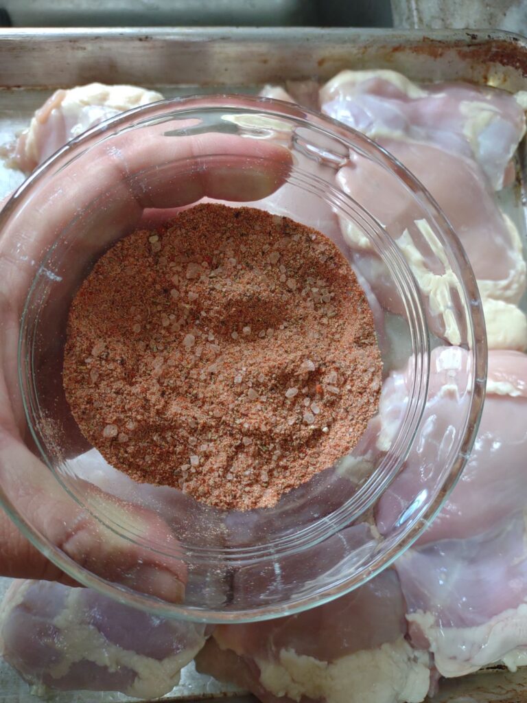 A dry rub in a glass bowl being held above raw chicken thighs.