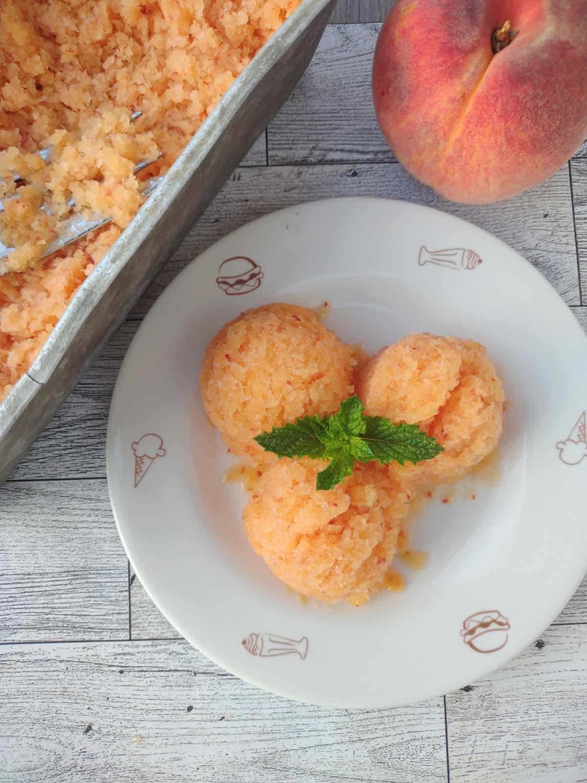 3 scoops of peach granita on a white plate with a peach and a metal loaf pan with more granita in it.