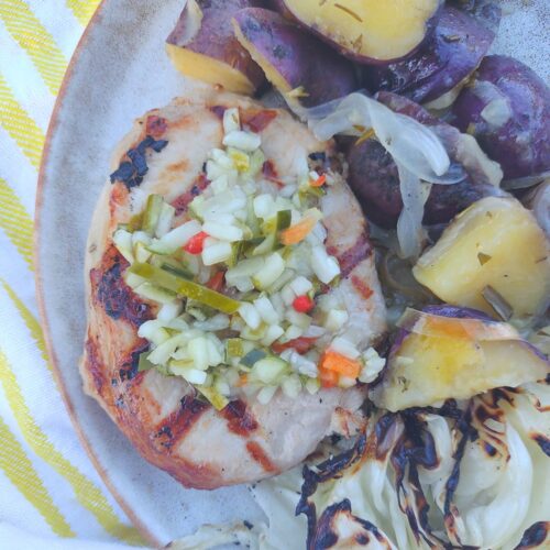 A pickle juice brined grilled pork chops topped with pickle de gallo on a plate with grilled cabbage and potatoes & onions.