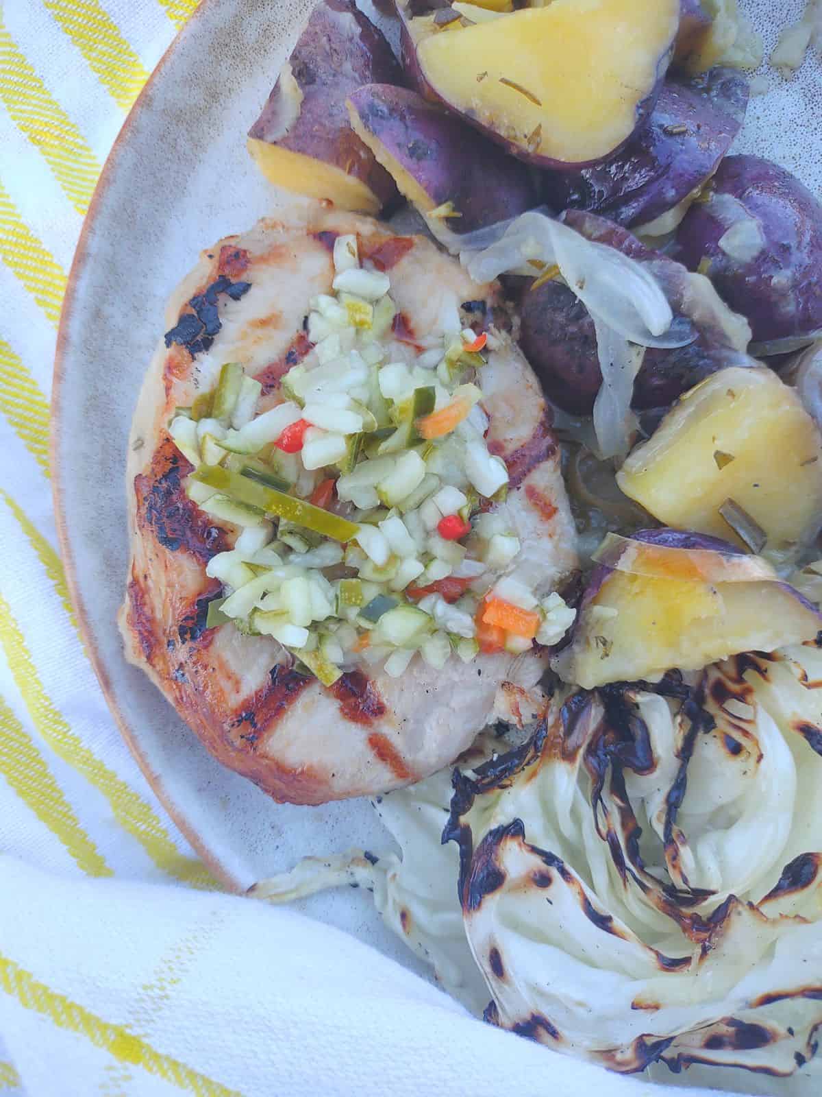 A pickle juice brined grilled pork chops topped with pickle de gallo on a plate with grilled cabbage and potatoes & onions.