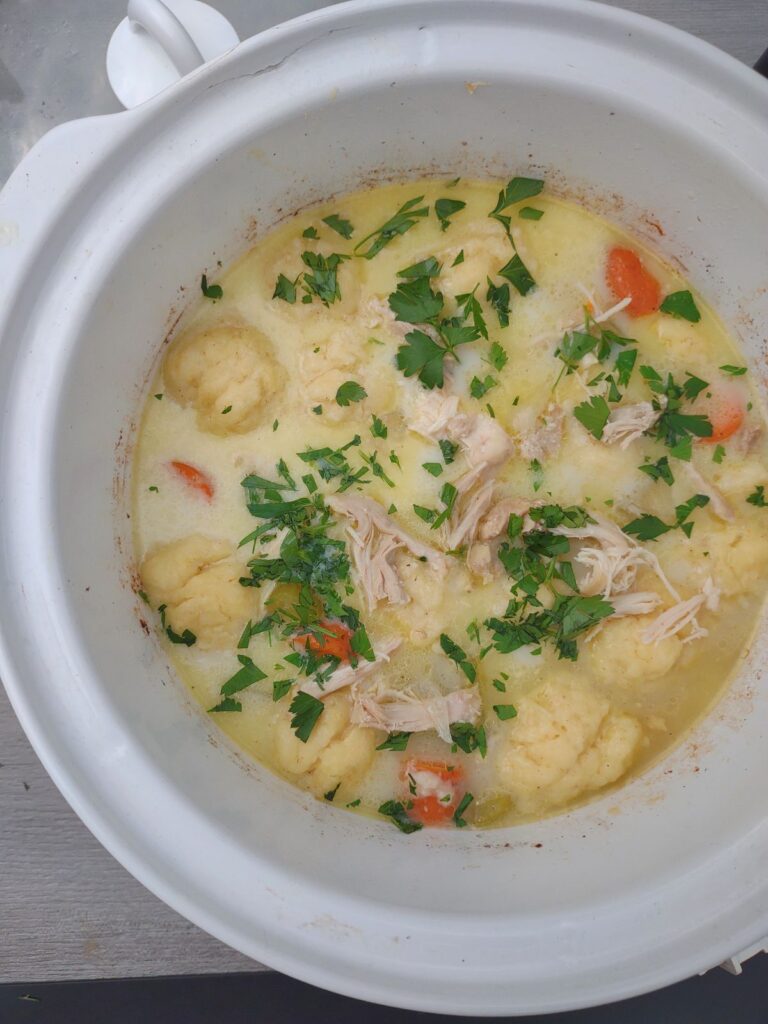 Chicken and dumplings with carrots, celery, and parsley in a white slow cooker.