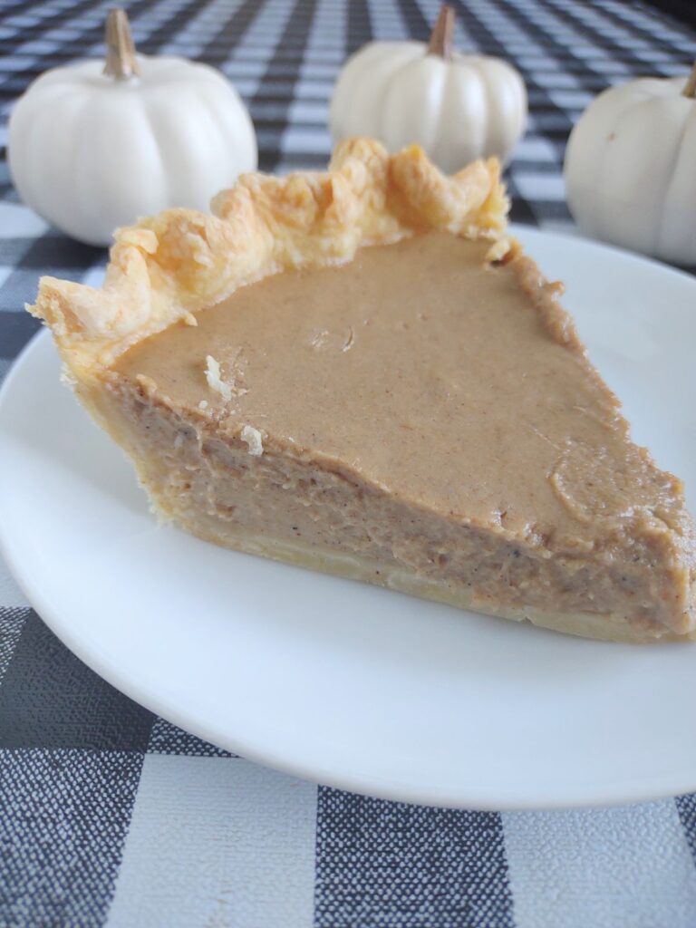 Creamy homemade pumpkin pie with flaky crust on a white plate, autumn decor with mini white pumpkins in background.