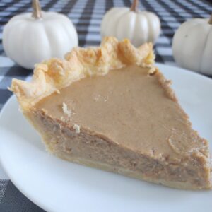 Creamy homemade pumpkin pie with flaky crust on a white plate, autumn decor with mini white pumpkins in background.
